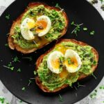 smashed avocado and boiled eggs on two slices of toast on a black plate with a microgreen garnish
